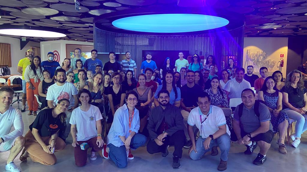 Grande grupo de pessoas sorrindo para a câmera, reunidas em um escritório moderno com iluminação colorida.