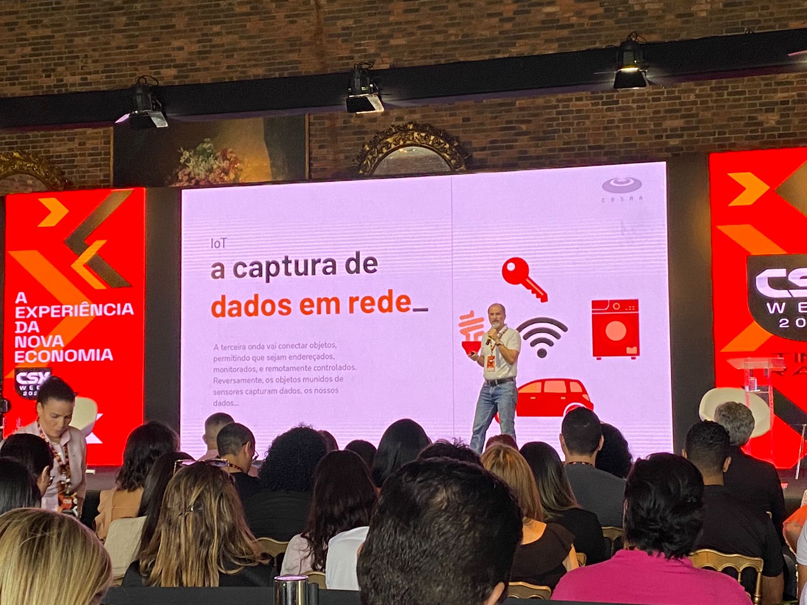 Um palestrante apresenta sobre a captura de dados em rede (IoT) em uma conferência, com uma tela mostrando ícones de dispositivos conectados e uma plateia atenta.