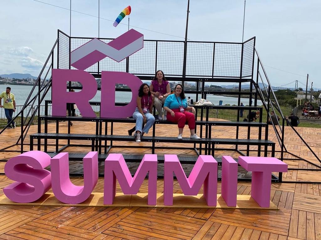 Três mulheres sentadas em um estande com as letras gigantes de "RD SUMMIT" na frente delas; um fundo de céu azul e água.