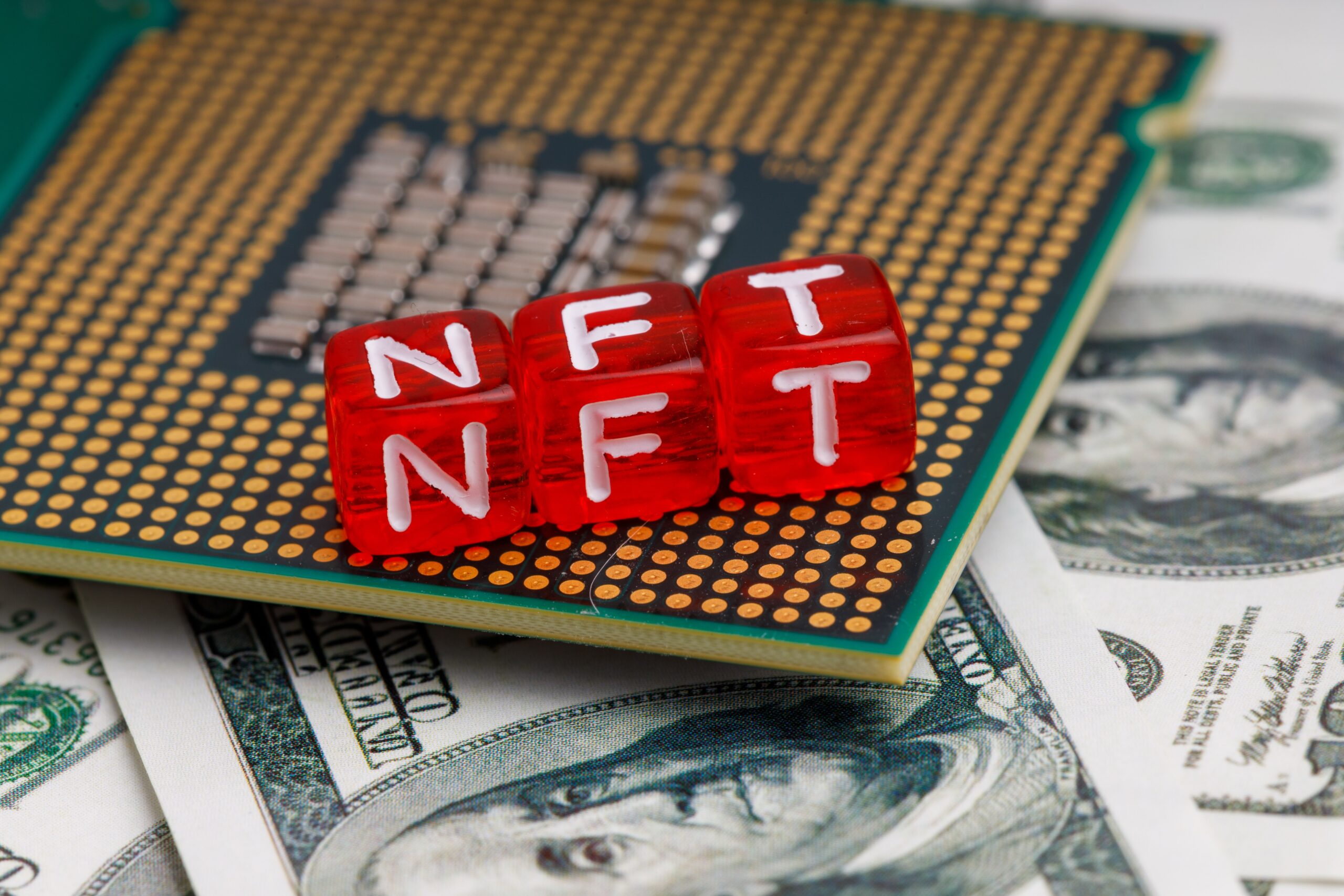 Três cubos vermelhos com a sigla "NFT" em branco sobre um processador de computador, apoiado em notas de dólar americano.