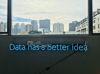Placa de néon azul com a frase "Data has a better idea" em uma parede de escritório com vista para uma cidade.