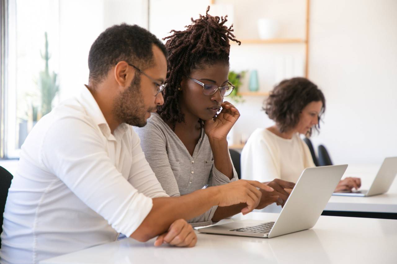 Dois colegas de trabalho, um homem e uma mulher, colaboram em um laptop, enquanto outra mulher trabalha em seu próprio laptop ao fundo.