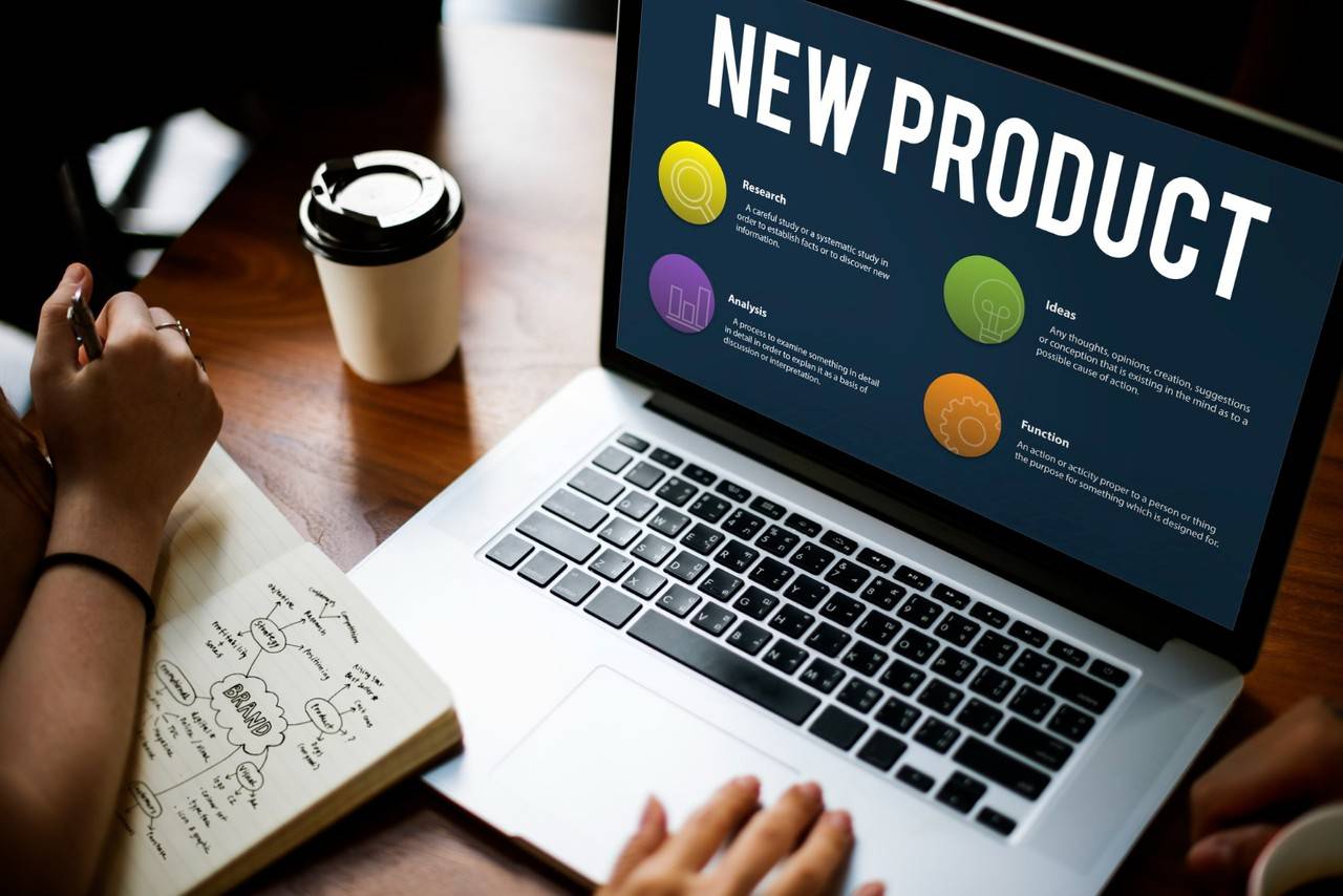 Laptop displaying "New Product" with sections on research, analysis, ideas, and function; a person takes notes nearby.