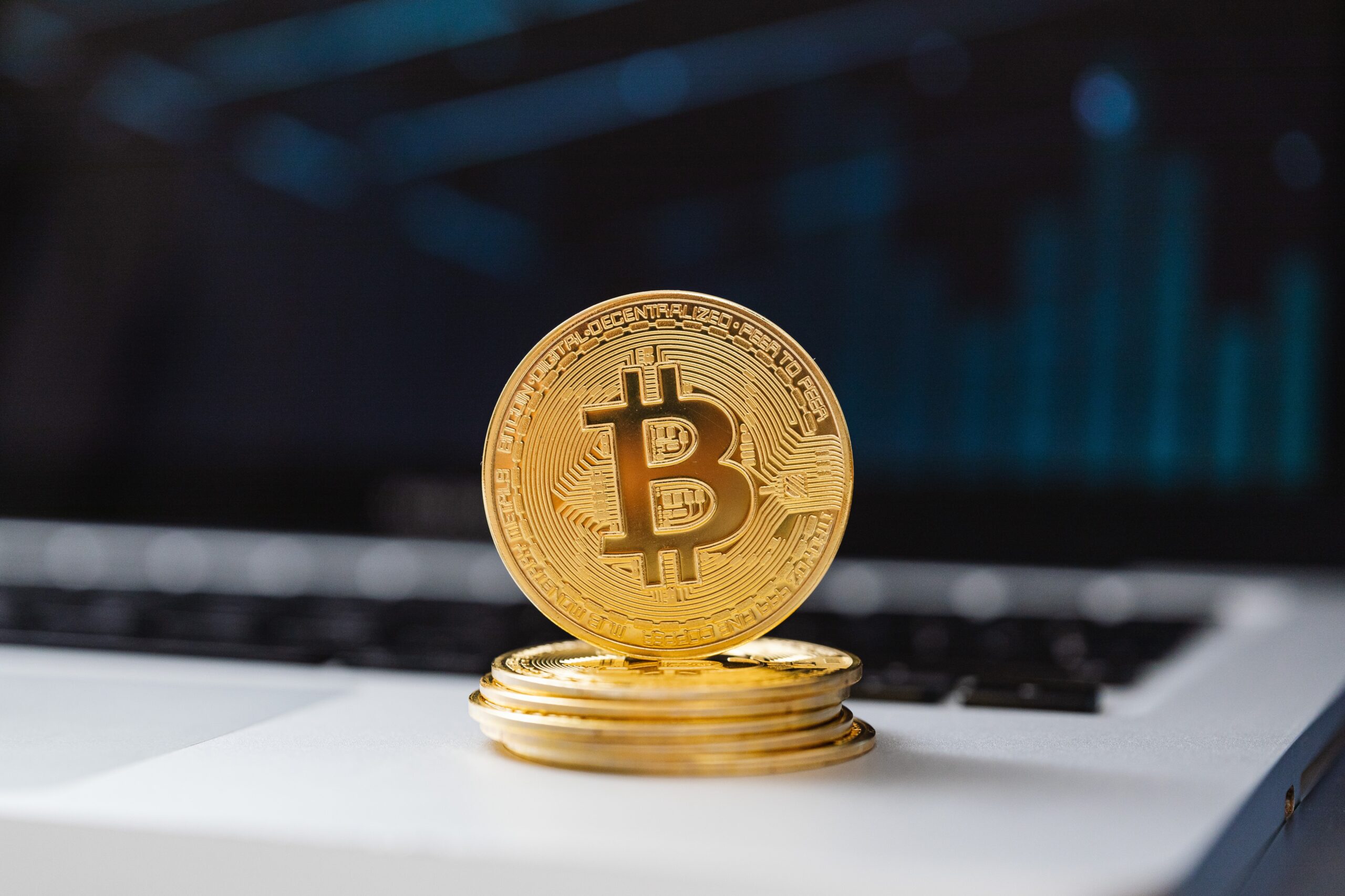 Uma pilha de bitcoins douradas em cima de um laptop, com um gráfico de ações desfocado ao fundo.