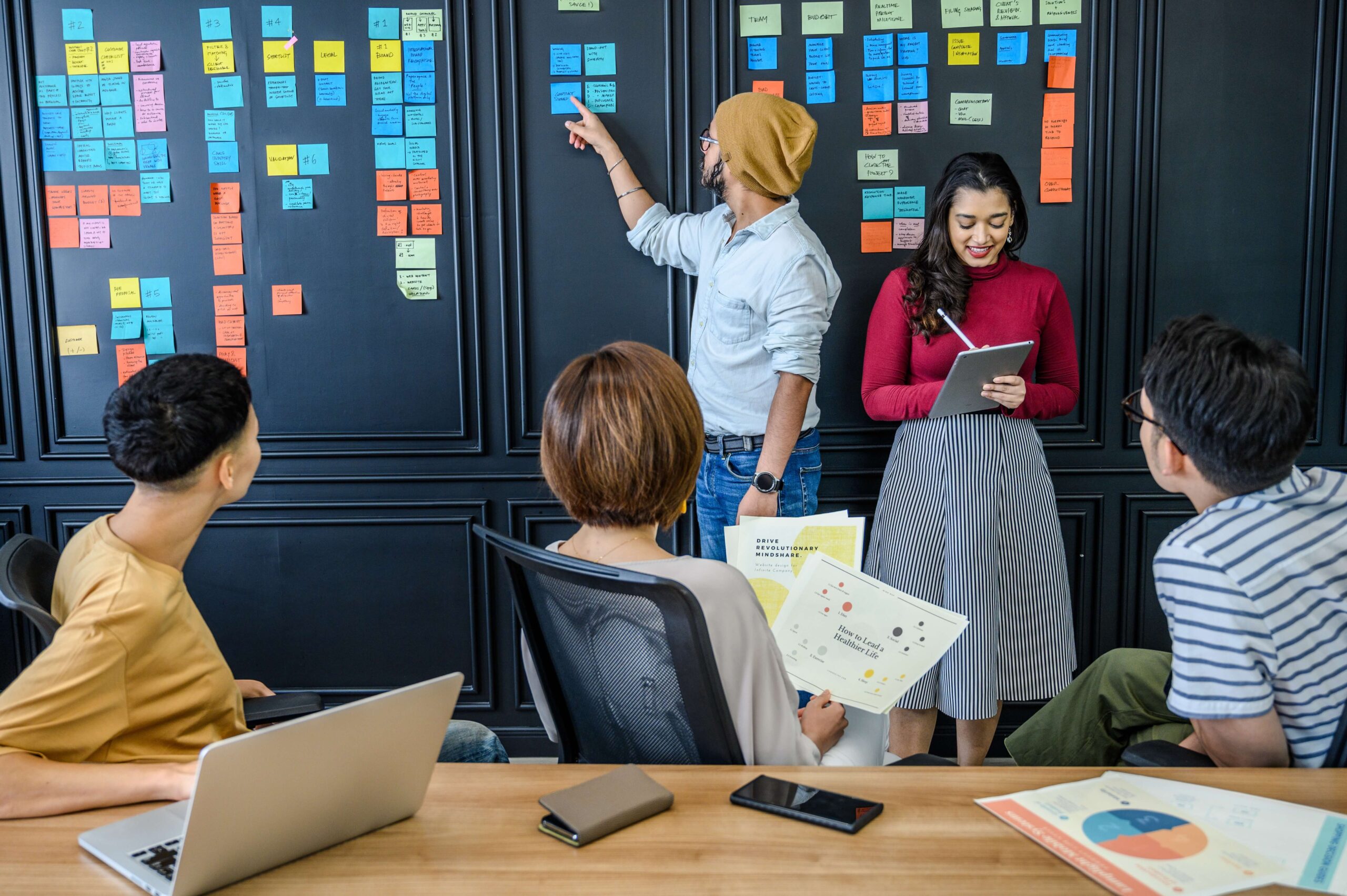 Um grupo de pessoas trabalha em conjunto numa sessão de brainstorming, com post-its coloridos num quadro negro e documentos gráficos espalhados numa mesa de conferência.