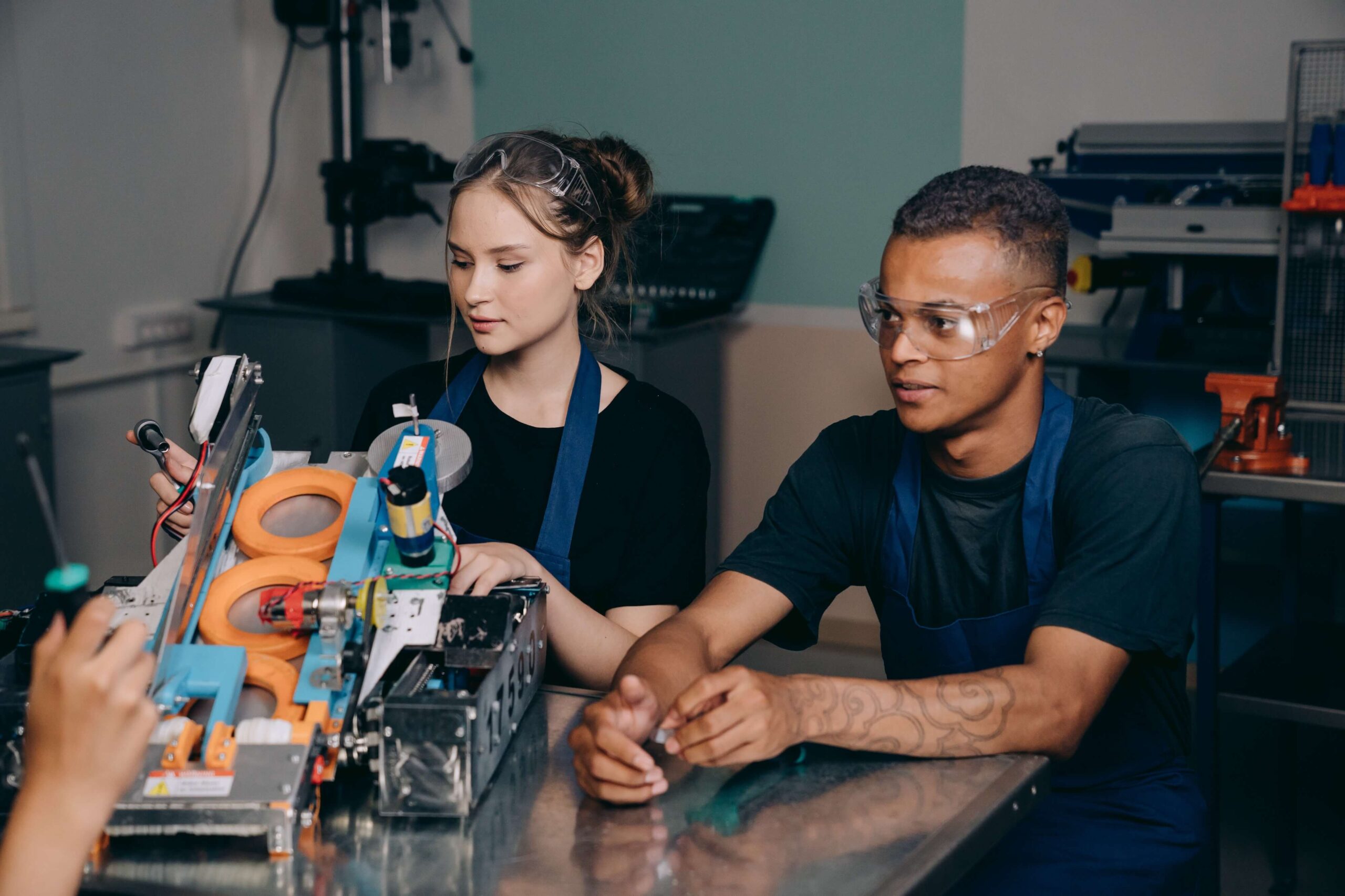 Dois jovens engenheiros trabalham juntos em um projeto de robótica numa oficina. Eles usam óculos de proteção e aventais enquanto inspecionam e manipulam um protótipo de máquina complexo.