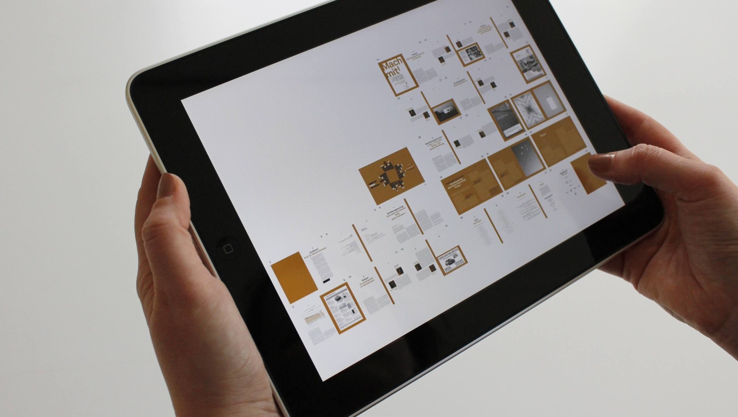 Duas mãos segurando um tablet que exibe uma publicação digital com páginas em miniatura em um layout de grade.