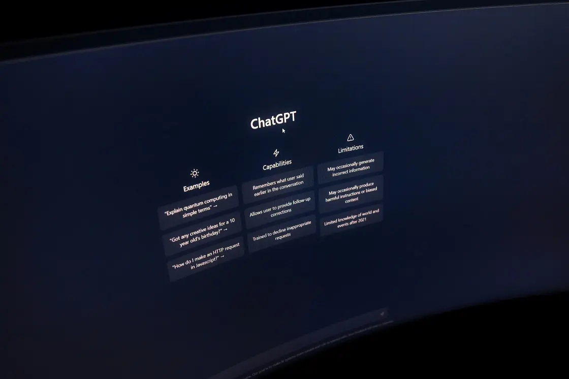 Monitor de computador exibindo a interface inicial do ChatGPT com colunas de exemplos, capacidades e limitações em fundo escuro.