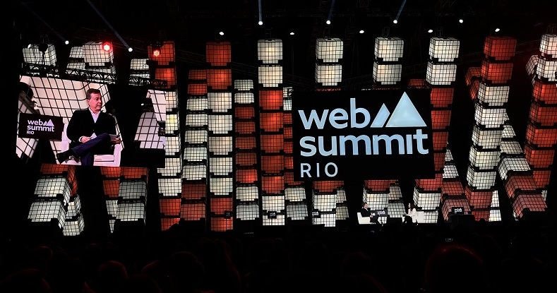 Um homem sentado em uma cadeira em um palco iluminado, com o logotipo da Web Summit Rio em destaque atrás dele, rodeado por uma estrutura de cubos iluminados em tons de branco e vermelho.