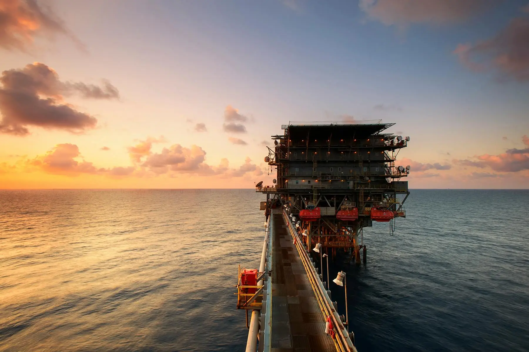 Plataforma de petróleo offshore conectada por passarela metálica sobre o mar durante o pôr do sol sob céu alaranjado.