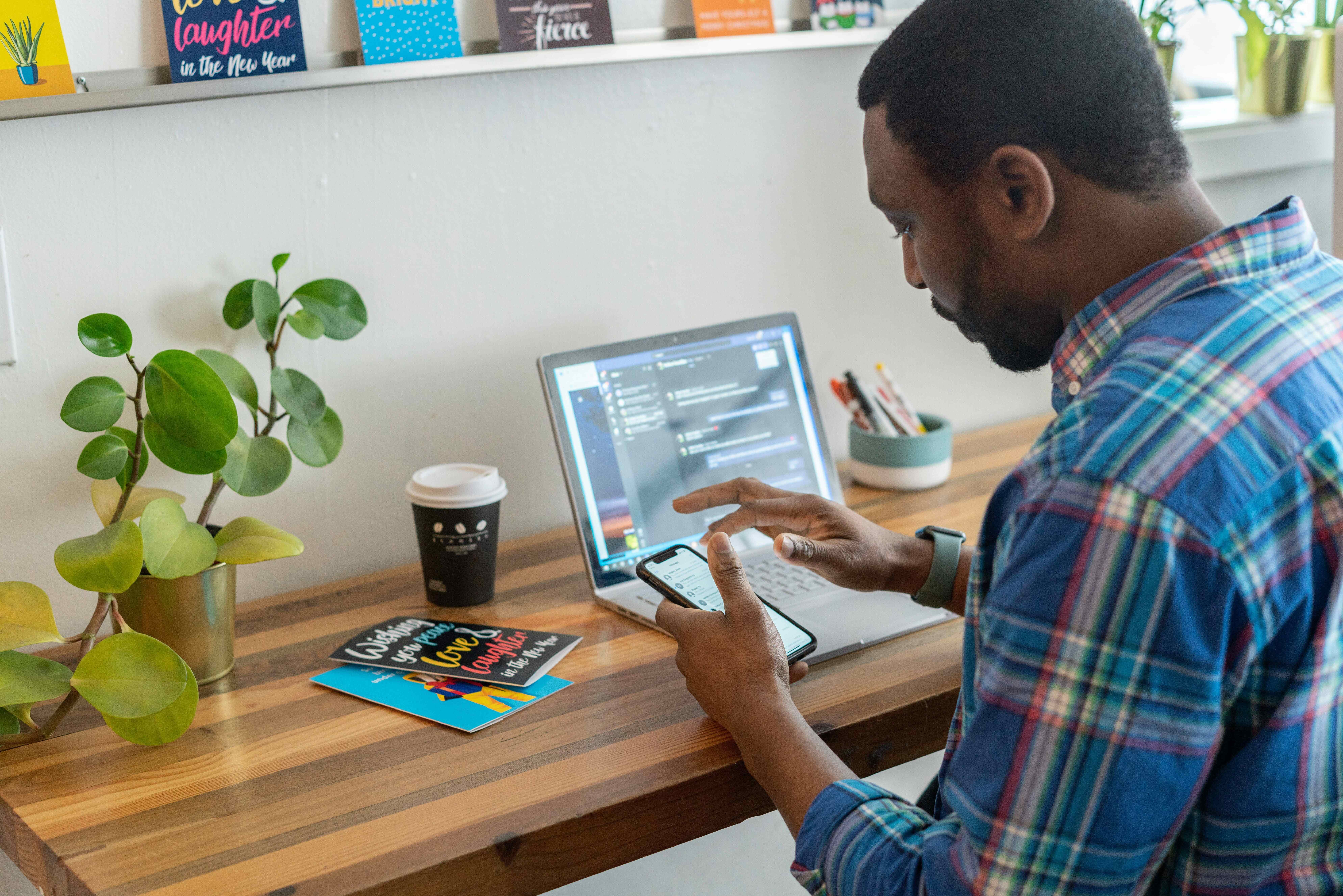 Home office scene: A man in a plaid shirt sits at a wooden desk, using his smartphone and laptop. Plants and greeting cards are visible on the desk.