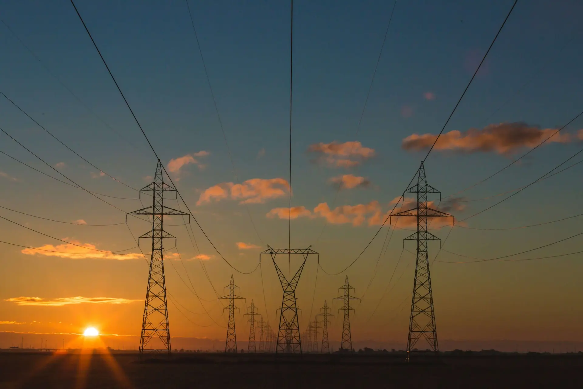 Torres de transmissão de energia elétrica em silhueta contra um pôr do sol alaranjado com céu azul e nuvens esparsas.