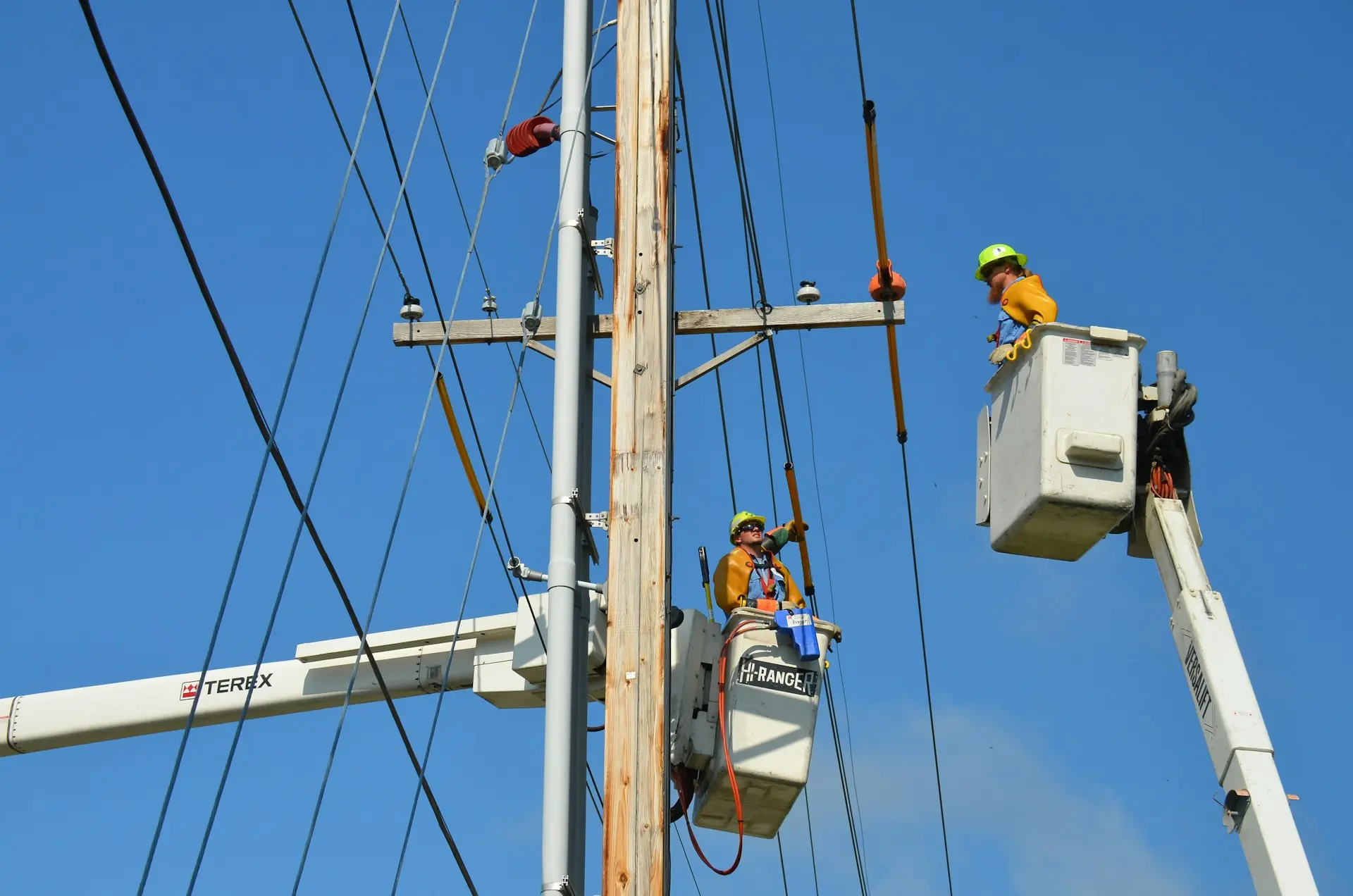Dois eletricistas em cestos aéreos de braços mecânicos realizam manutenção em fiação de postes elétricos sob céu azul.