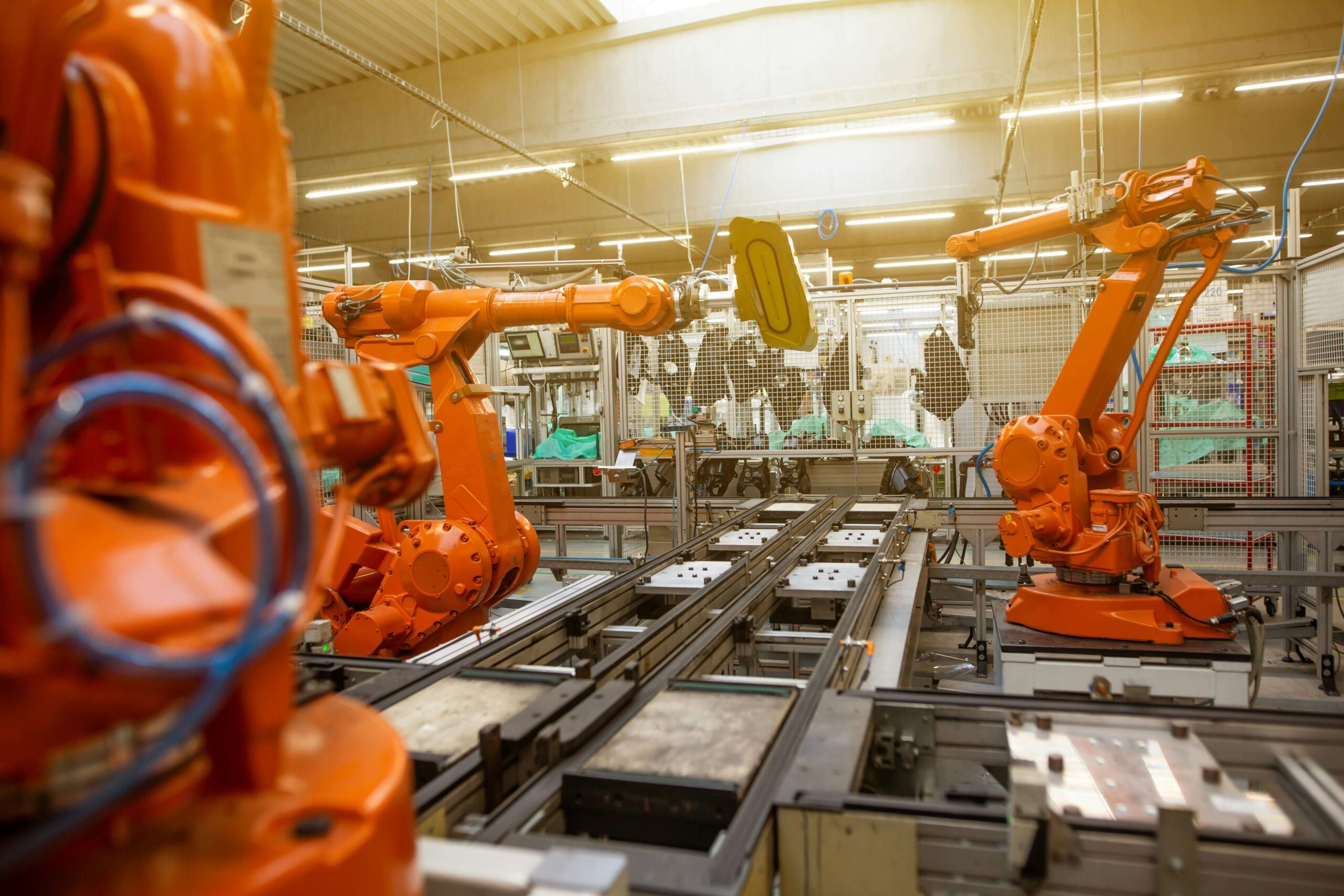 Braços robóticos laranja em uma linha de montagem industrial moderna, movendo peças em uma esteira transportadora.