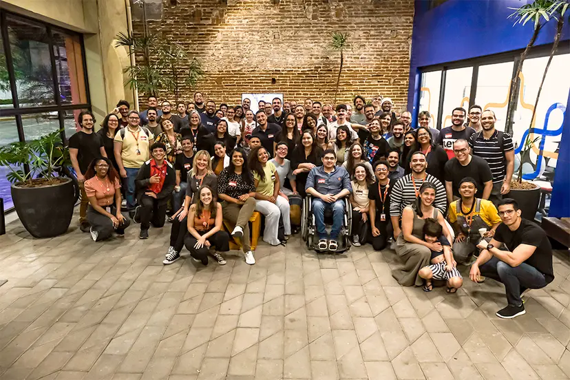 Grupo numeroso de alunos e mentores sorrindo em um pátio com parede de tijolos.