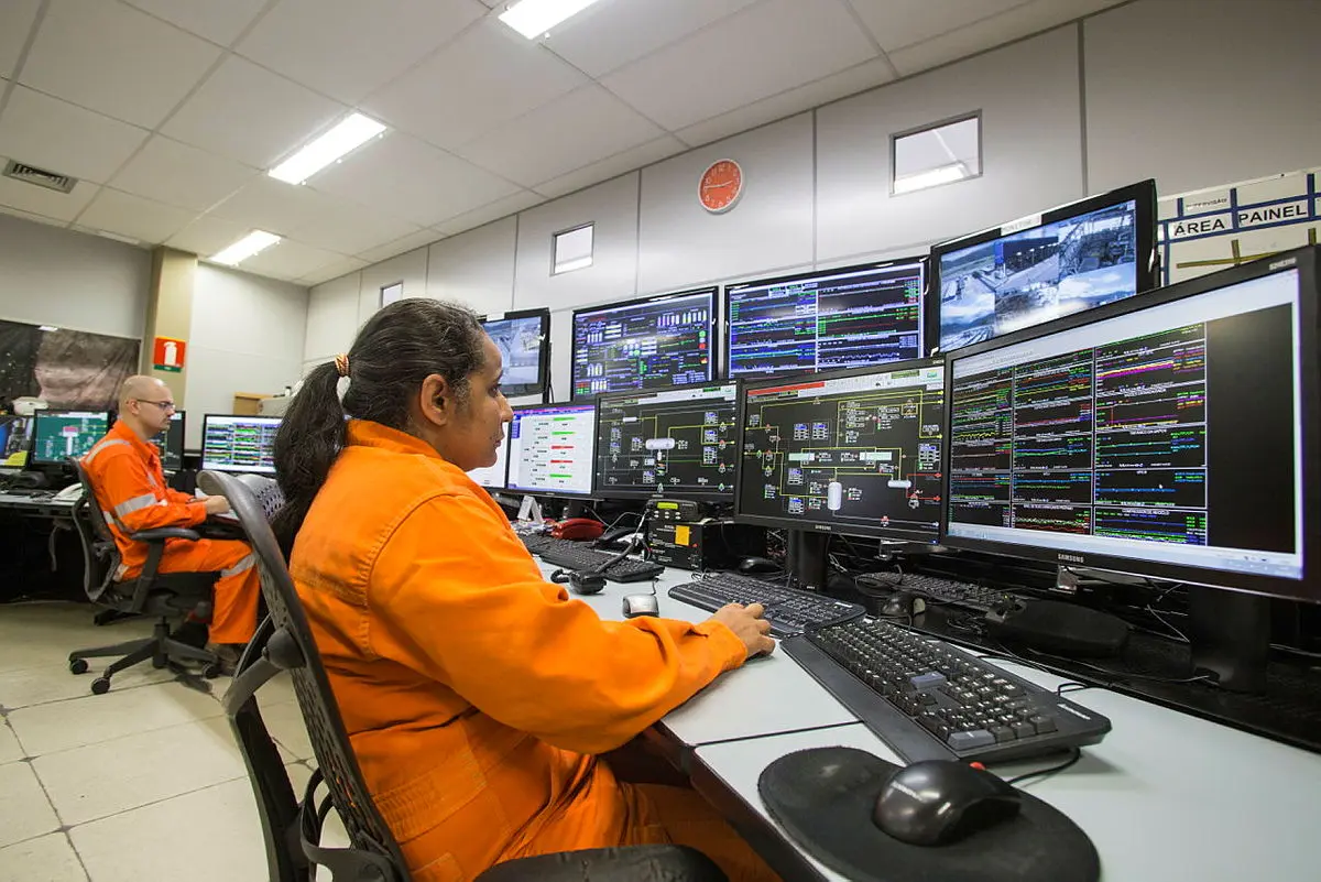 Mulher e homem em macacão laranja operam computadores com múltiplos ecrãs de gráficos e mapas num centro de controlo de energia.