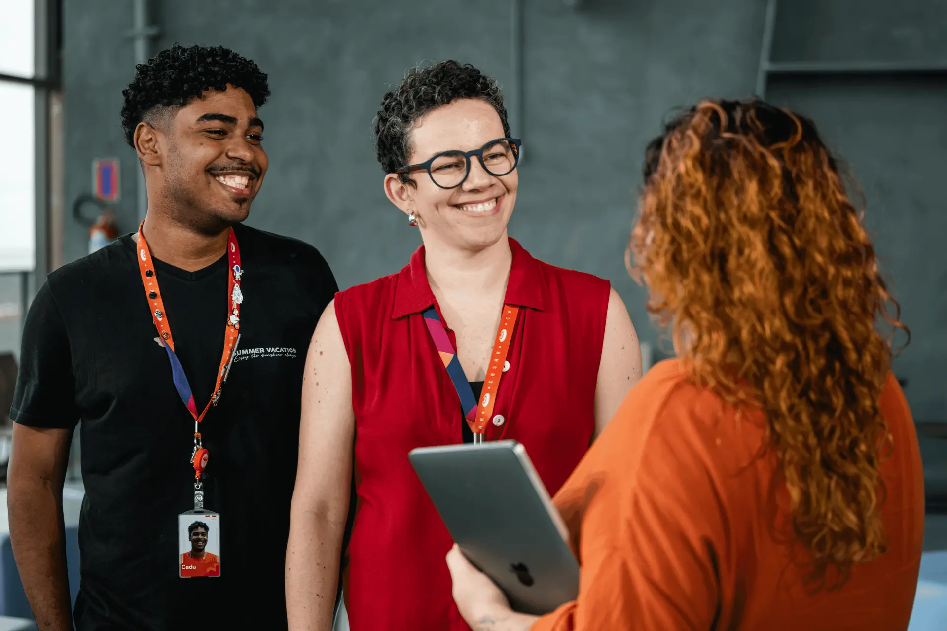 Três pessoas sorrindo em uma conversa de trabalho; um homem e uma mulher de crachá ouvem uma colega que segura um tablet.