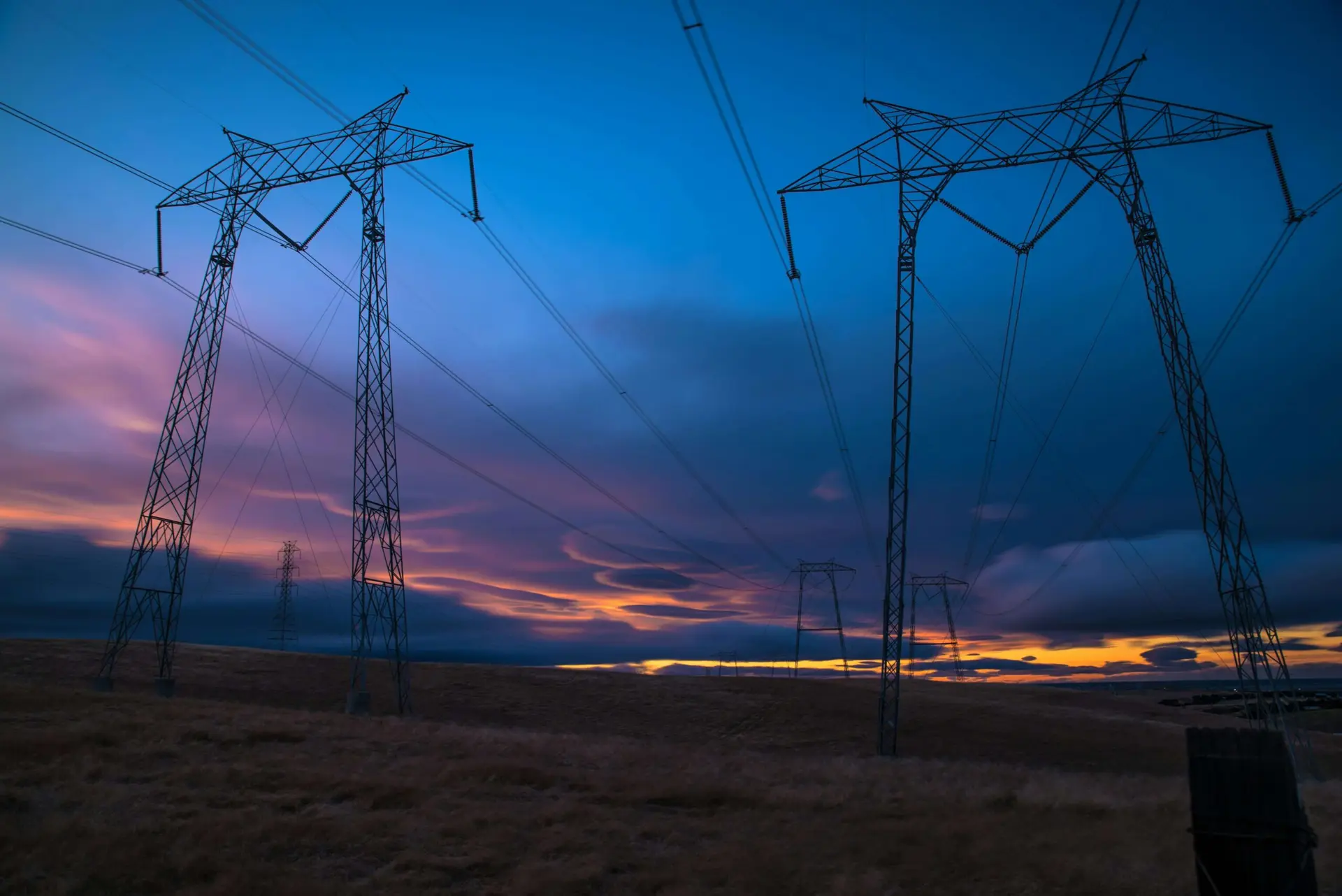 Duas grandes torres de transmissão de energia em um campo aberto sob céu ao entardecer com tons de azul, rosa e laranja.