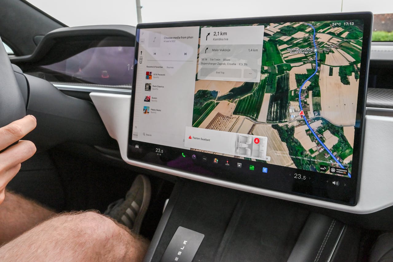 Close-up of a Tesla's central touchscreen displaying navigation, music options, and a seatbelt reminder; driver's legs and hand on the steering wheel are partially visible.