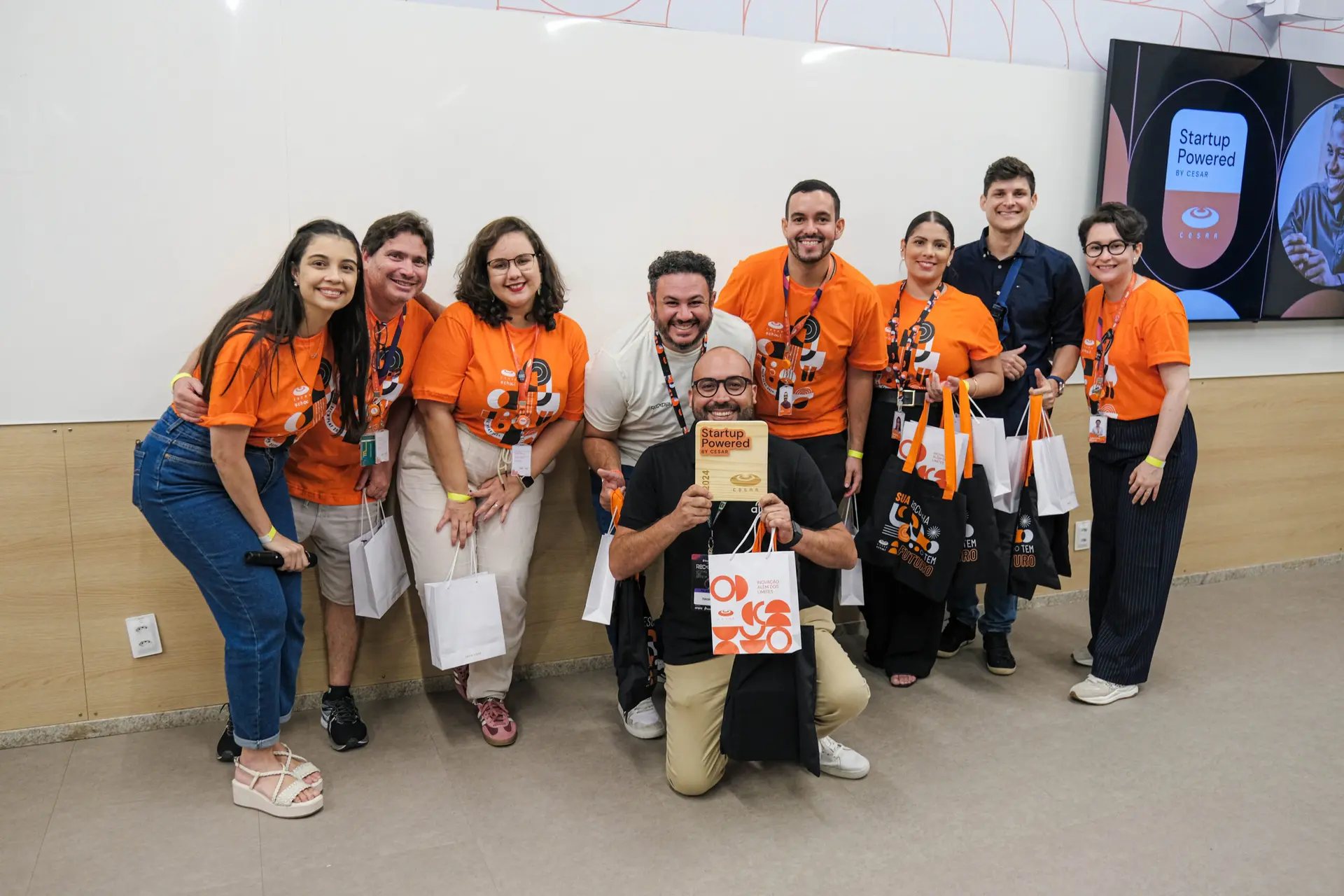 Grupo de nove pessoas sorrindo; homem central ajoelhado segura placa "Startup Powered by CESAR" em ambiente de evento de inovação.