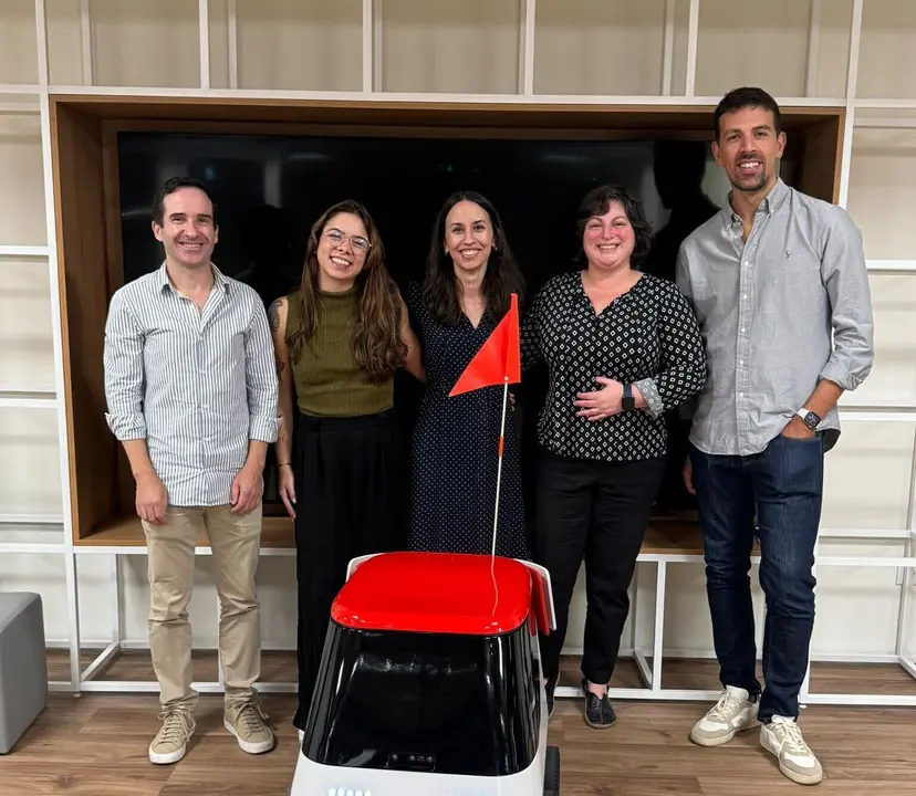 Cinco profissionais sorridentes posam atrás de um pequeno robô de entregas branco e vermelho com uma bandeira laranja.