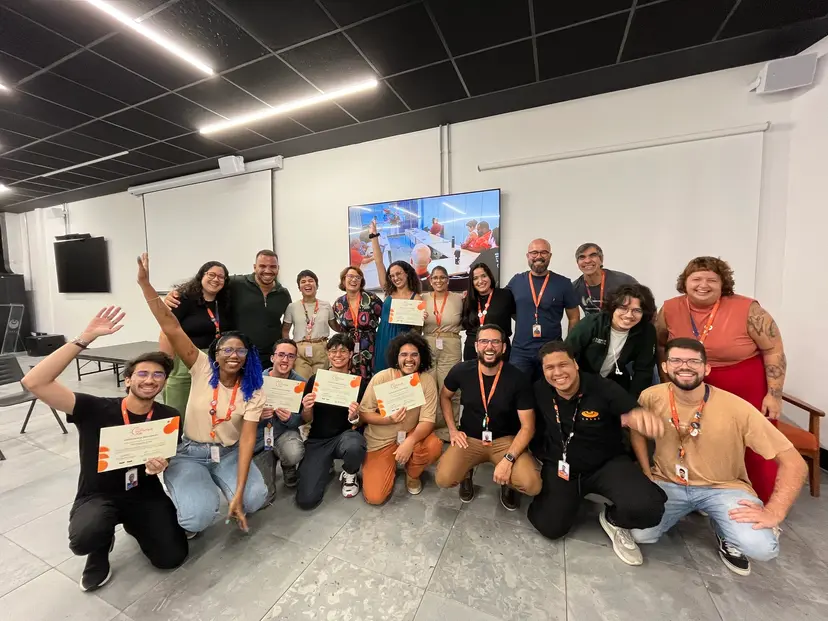 Grupo de 18 profissionais sorridentes segura certificados em sala de treinamento, celebrando a conclusão de projeto de inovação