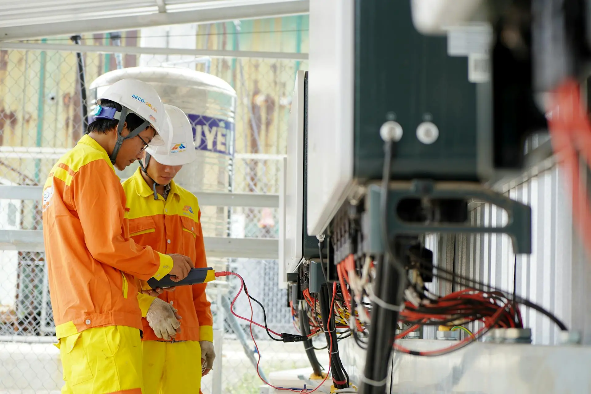 Dois técnicos de capacete e uniforme laranja realizam testes elétricos em um painel de controle usando um multímetro digital.