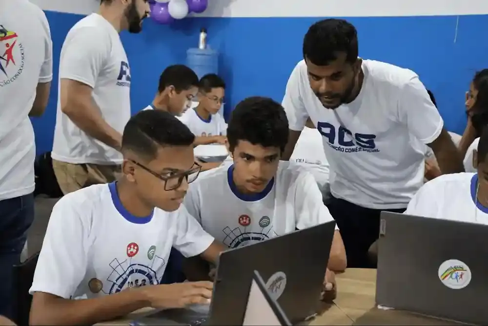 Três jovens concentrados utilizam computadores portáteis enquanto um instrutor orienta a atividade em sala de aula do CESAR.