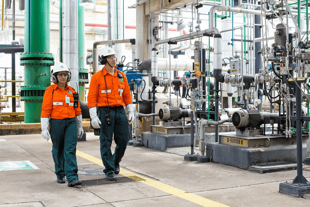 Dois colaboradores caminham por uma planta industrial com tubulações e equipamentos mecânicos enquanto realizam verificação de sobressalentes, usando capacetes, protetores auriculares, óculos de segurança e uniforme