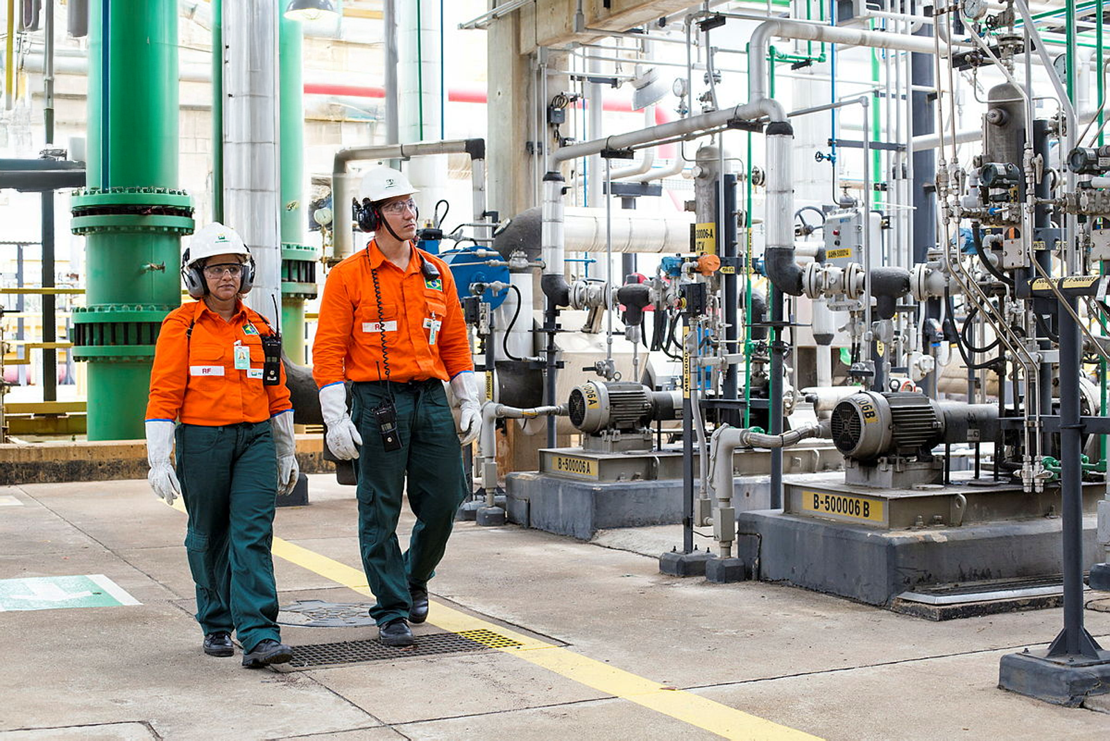 Dois colaboradores caminham por uma planta industrial com tubulações e equipamentos mecânicos enquanto realizam verificação de sobressalentes, usando capacetes, protetores auriculares, óculos de segurança e uniforme
