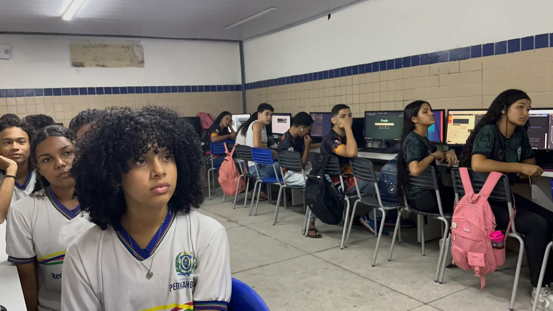 Grupo de jovens em laboratório de informática escolar, sentados em frente a computadores de mesa, usando uniformes de Pernambuco.