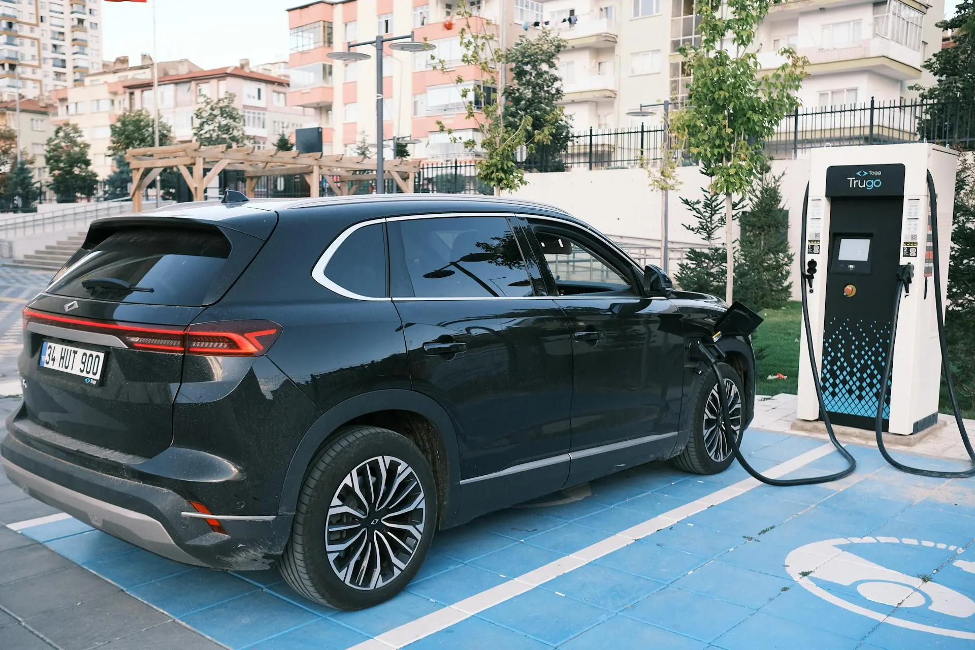 SUV preto estacionado em vaga azul enquanto carrega em totem de energia elétrica em ambiente urbano com prédios ao fundo.