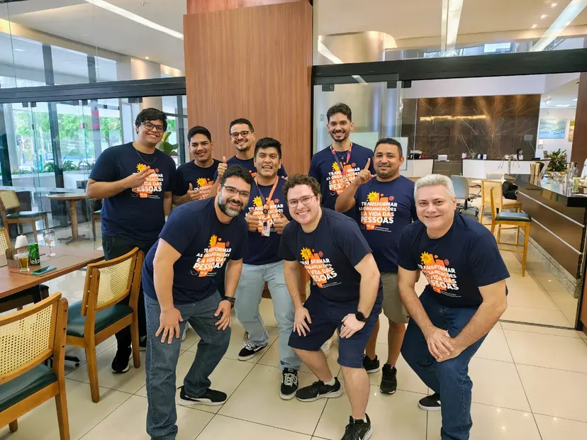 Nove homens sorridentes com t-shirts azuis iguais posam num restaurante. A frase na camisa foca na transformação de pessoas.
