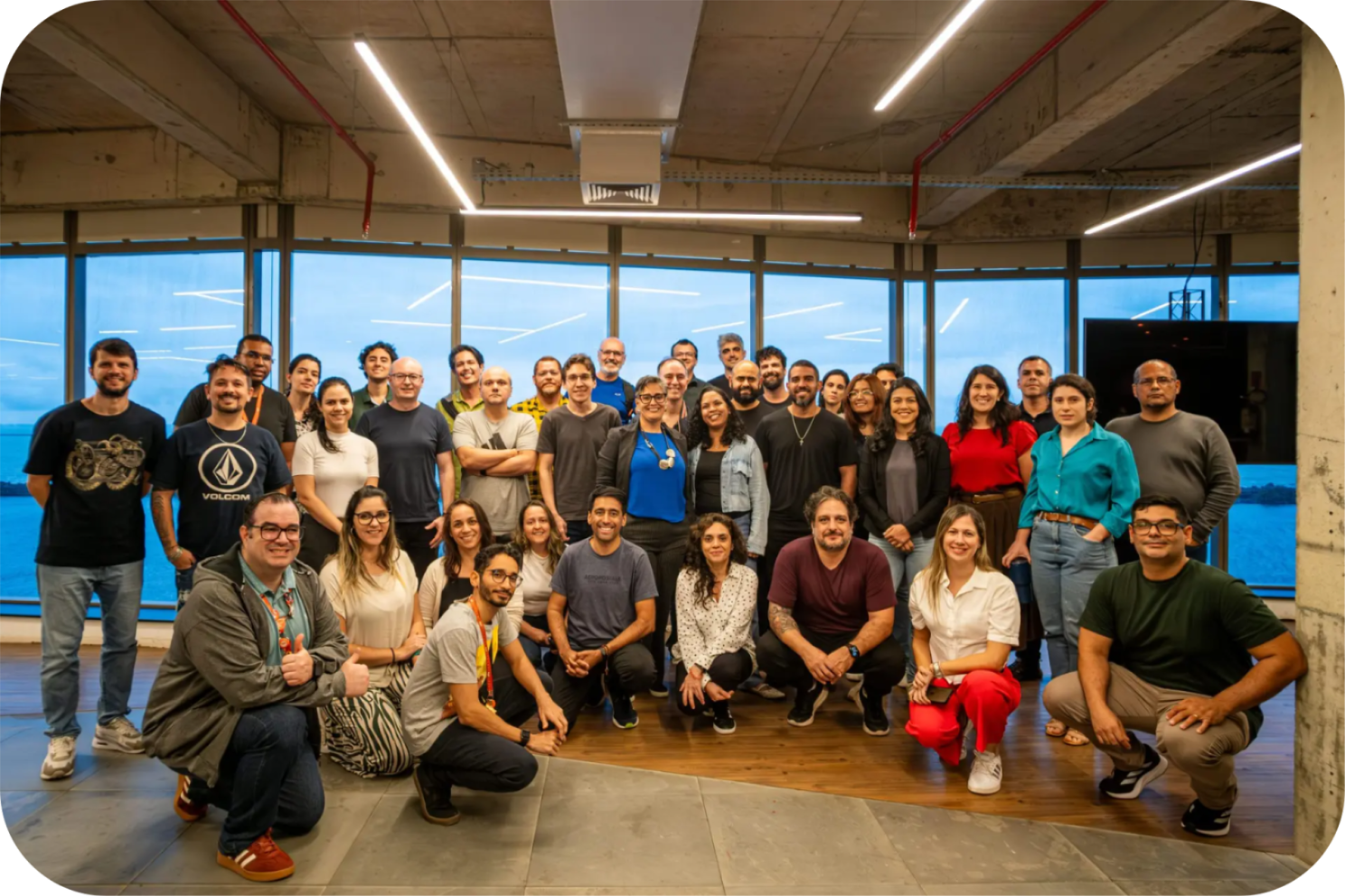 Grupo de cerca de 30 profissionais sorridentes posa em escritório moderno com grandes janelas de vidro ao fundo.