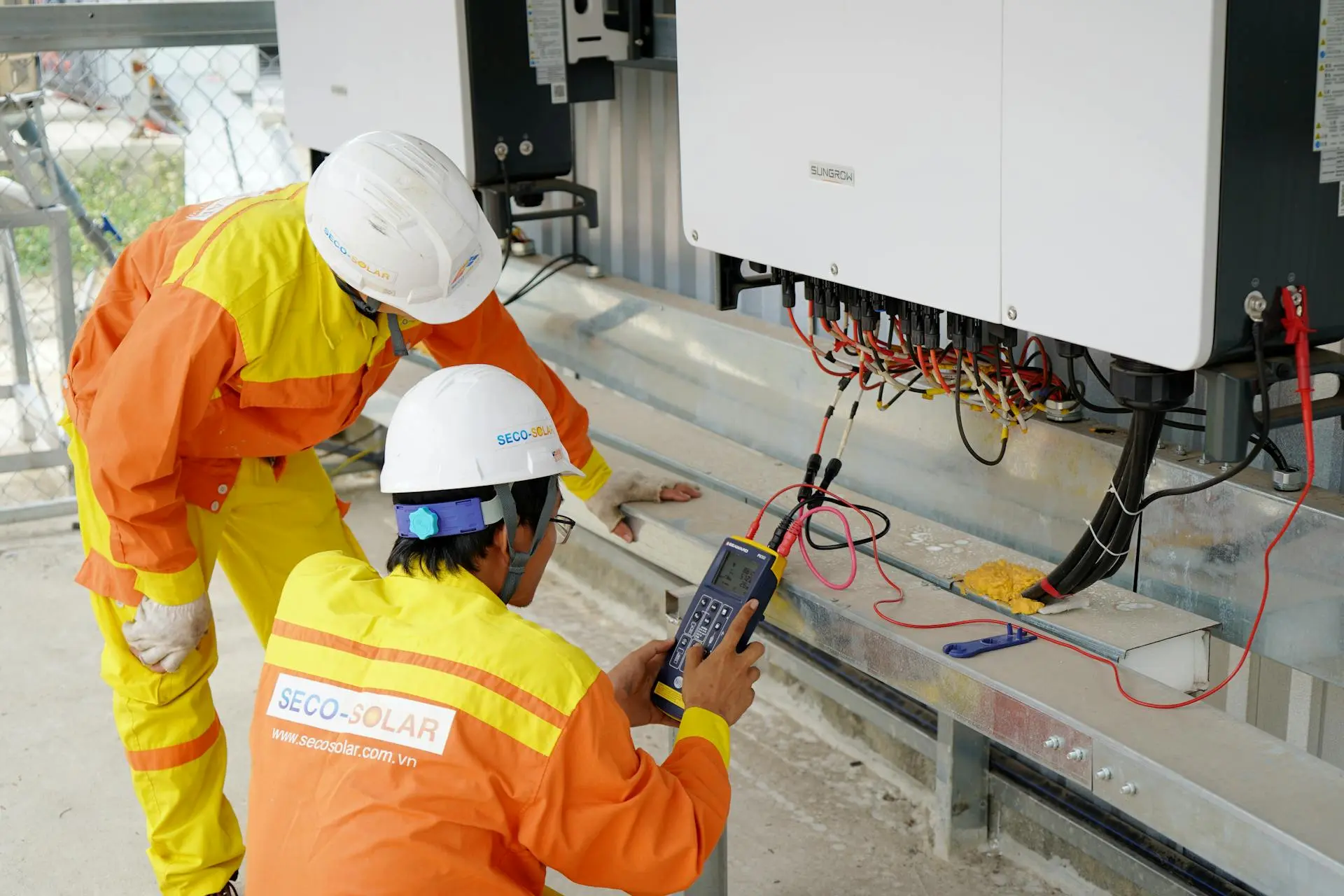 Dois técnicos de uniforme laranja e capacete branco utilizam um medidor digital em painel de sistema de energia solar.