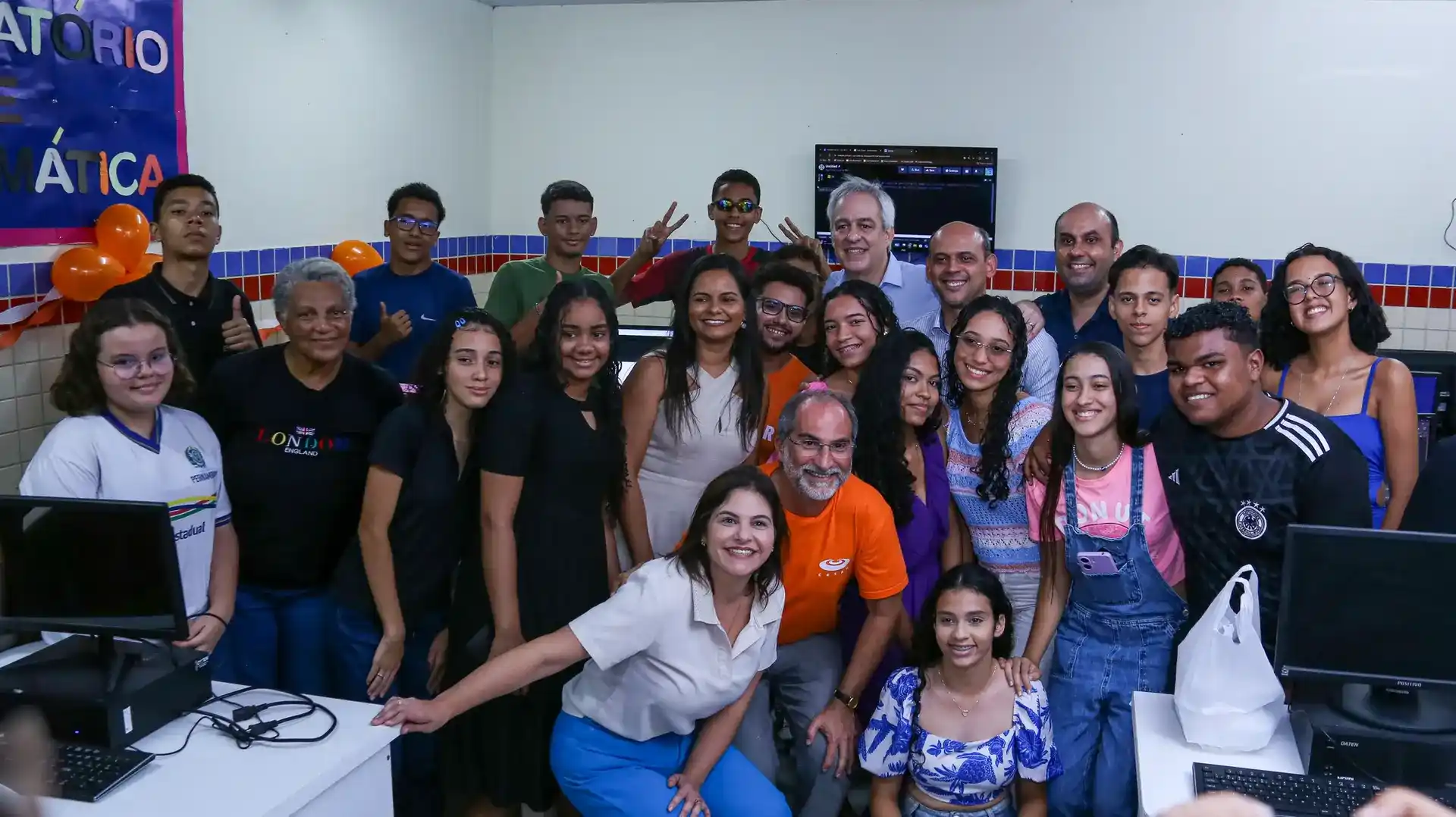 Grupo de alunos e educadores sorridentes em laboratório de informática escolar com computadores ao fundo