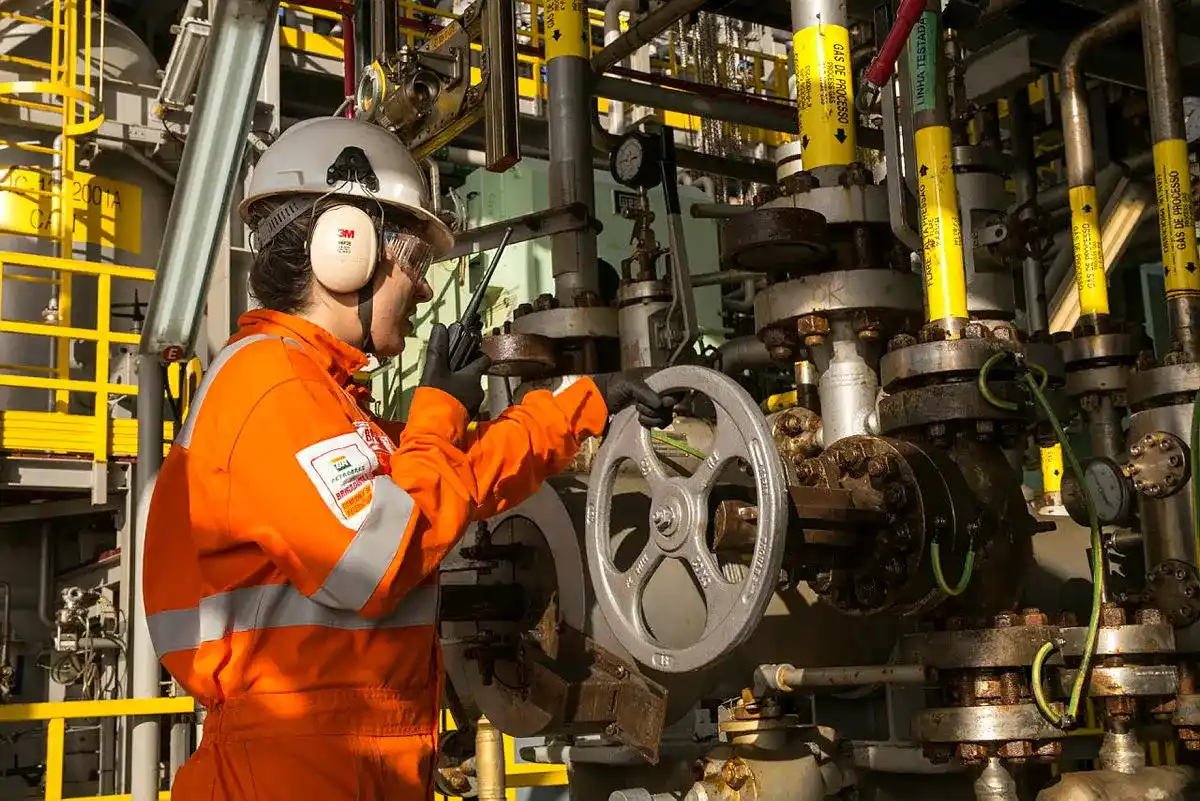 Engenheira com macacão laranja, capacete e rádio, opera válvula em complexa infraestrutura de tubagens