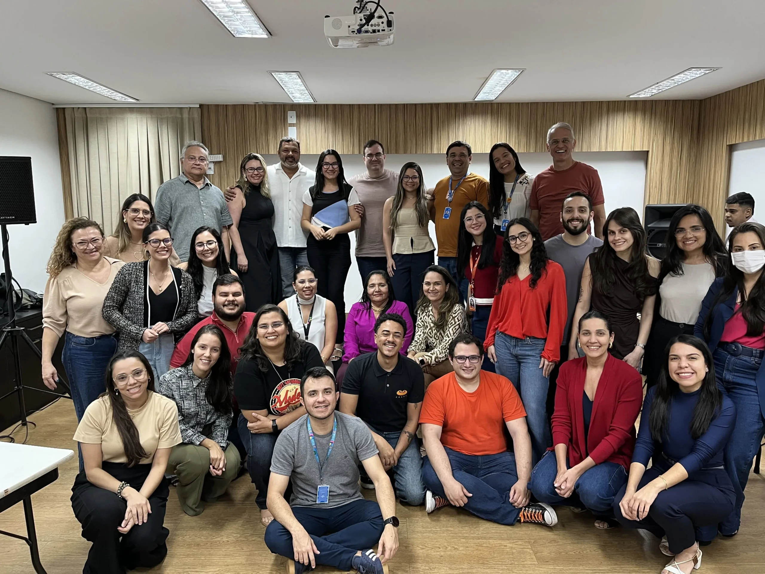 Grupo de aproximadamente 30 profissionais sorrindo em uma sala de treinamento. Estão distribuídos em pé, sentados e agachados