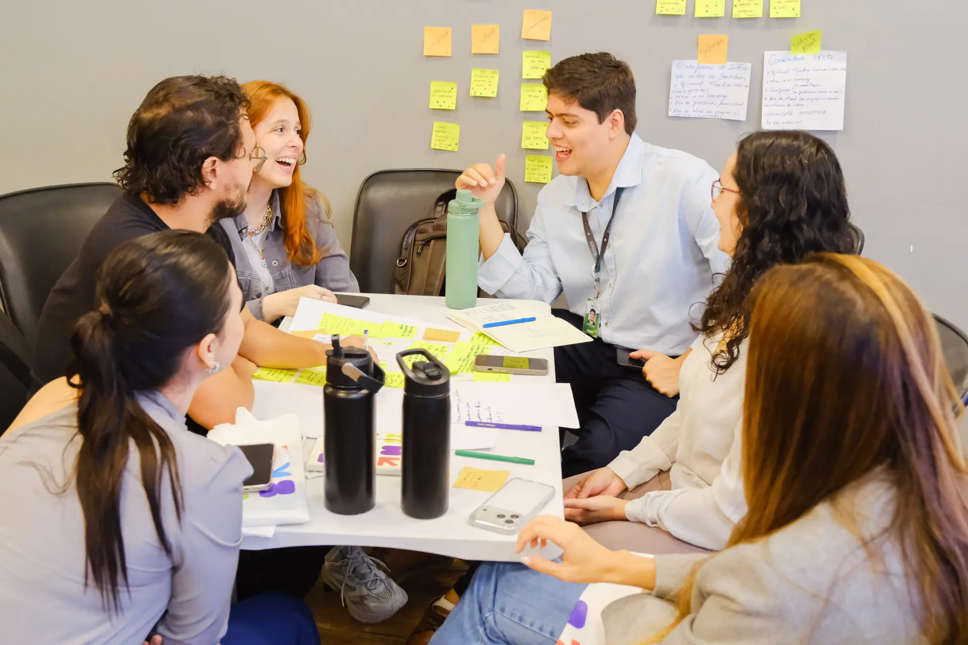 Seis pessoas em reunião de inovação corporativa fazem brainstorming com post-its, anotações na parede e materiais sobre a mesa