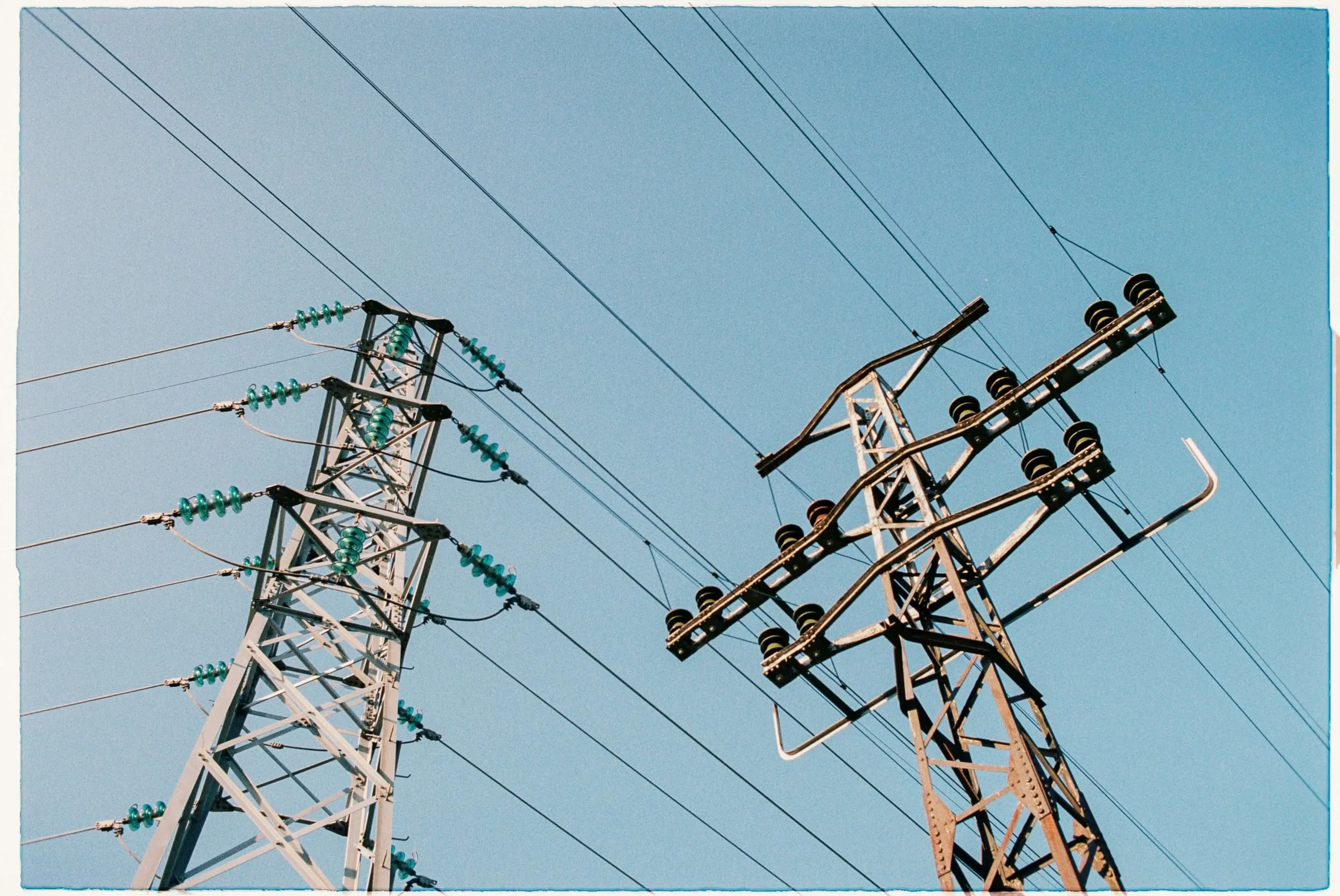 Duas torres de transmissão de energia de metal sob céu azul claro, destacando cabos de alta tensão e isoladores de cerâmica.
