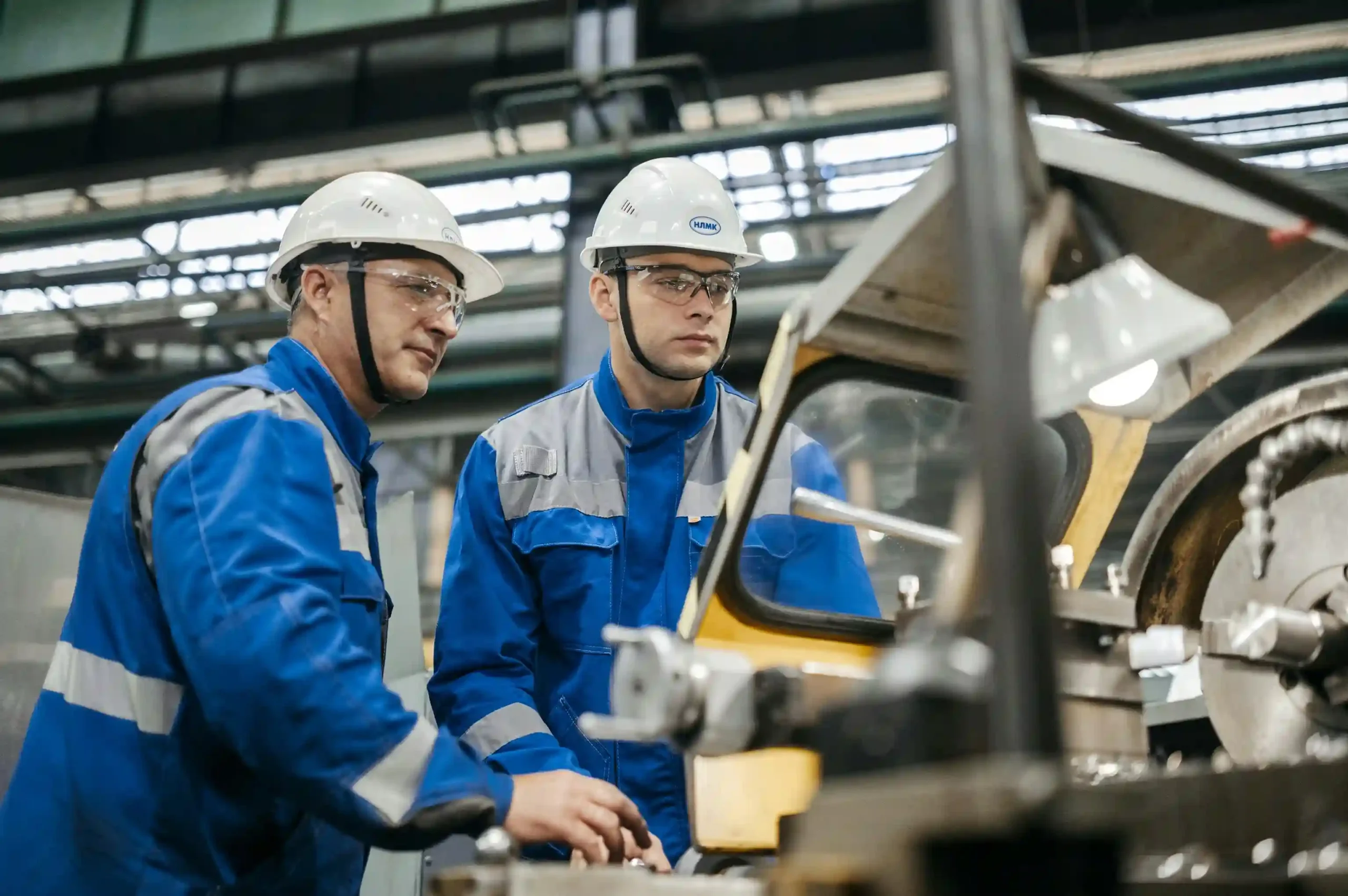 Dois operários de uniforme azul, capacete branco e óculos de proteção operam maquinário pesado em ambiente de fábrica industrial.
