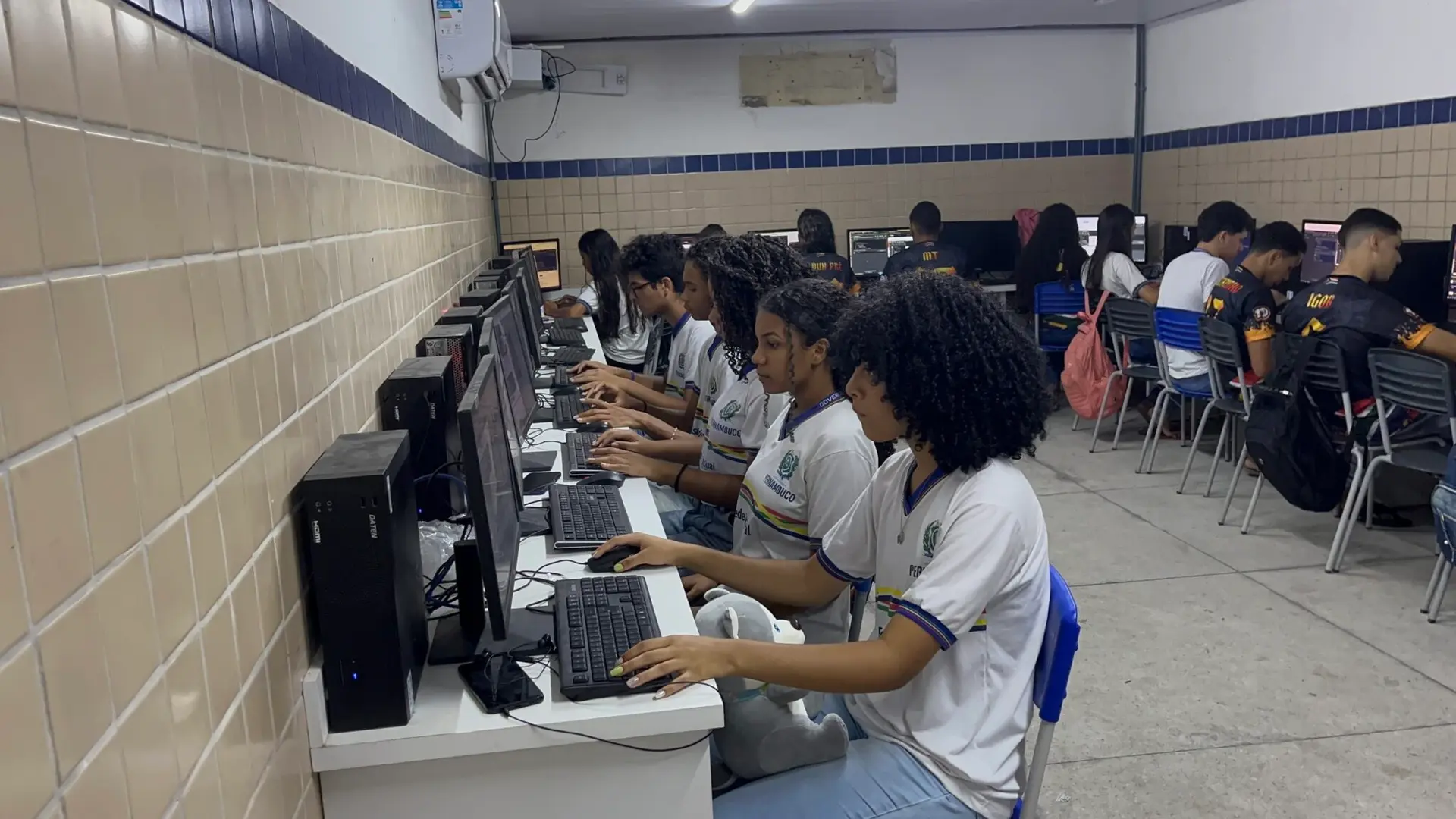 Grupo de jovens sentados em bancadas usando computadores desktop em um laboratório de informática escolar com paredes de azulejo.