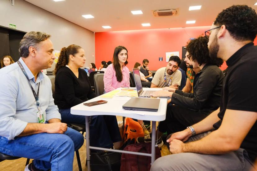 Educação Corporativa Integrada à Tecnologia e Estratégia para Resultados Grupo de seis profissionais reunidos em torno de uma mesa branca com notebook e post-its em sala de treinamento moderna.
