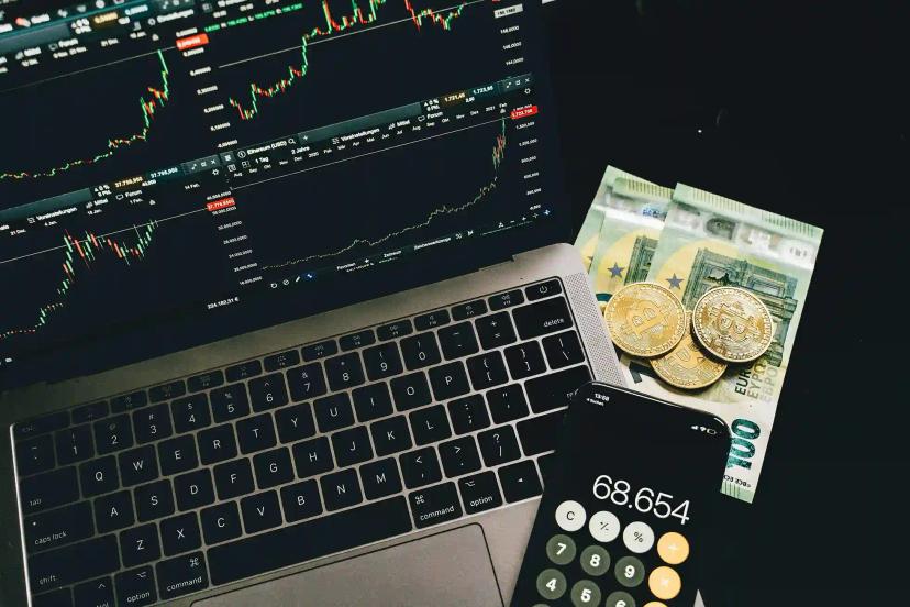 IA generativa no setor financeiro Portátil exibe gráficos financeiros, ao lado de notas de euro, moedas de Bitcoin e um telemóvel com calculadora aberta.