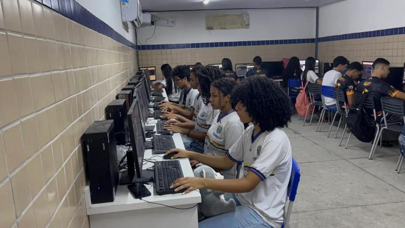Impacto Social na Educação e Conexão de Estudantes ao Mercado Tech Grupo de jovens sentados em bancadas usando computadores desktop em um laboratório de informática escolar com paredes de azulejo.