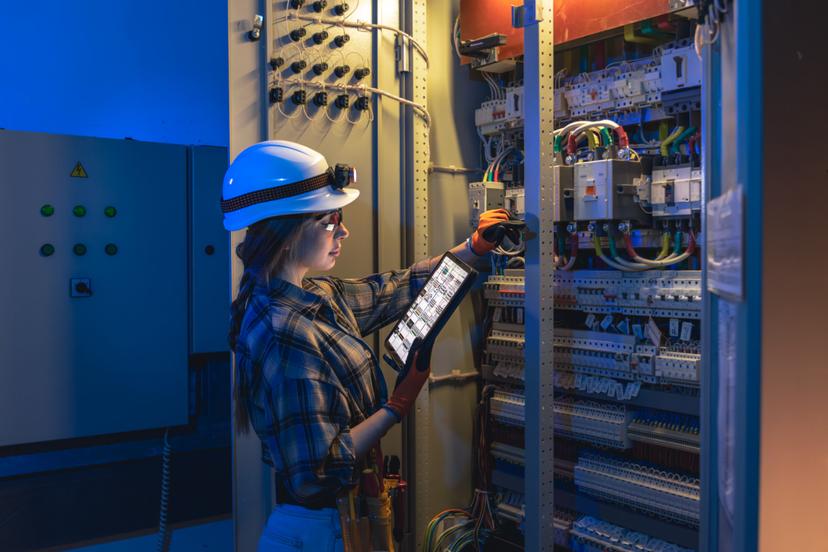 Governança de IA no setor de energia eletrica Eletricista de capacete branco e óculos de proteção utiliza tablet para inspecionar fiação e disjuntores em painel elétrico.